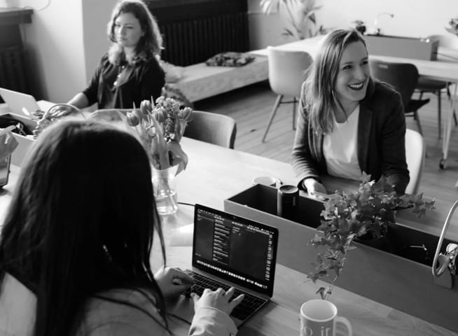 Three women work at the desk in their office, one sitting on her back, the other two facing her. One of them laughs as she talks to someone out of the camera frame